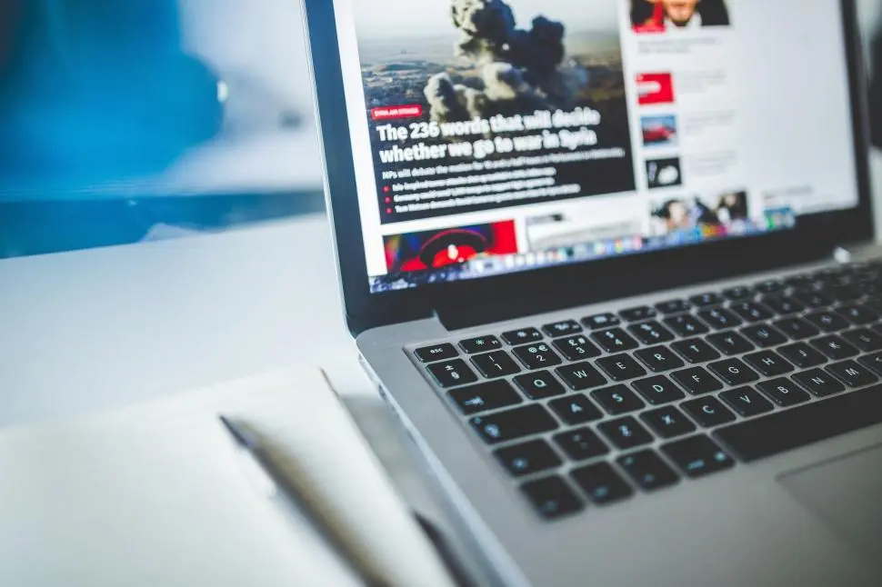 Close-up of a laptop displaying a news website, with a notebook and pen placed beside it.